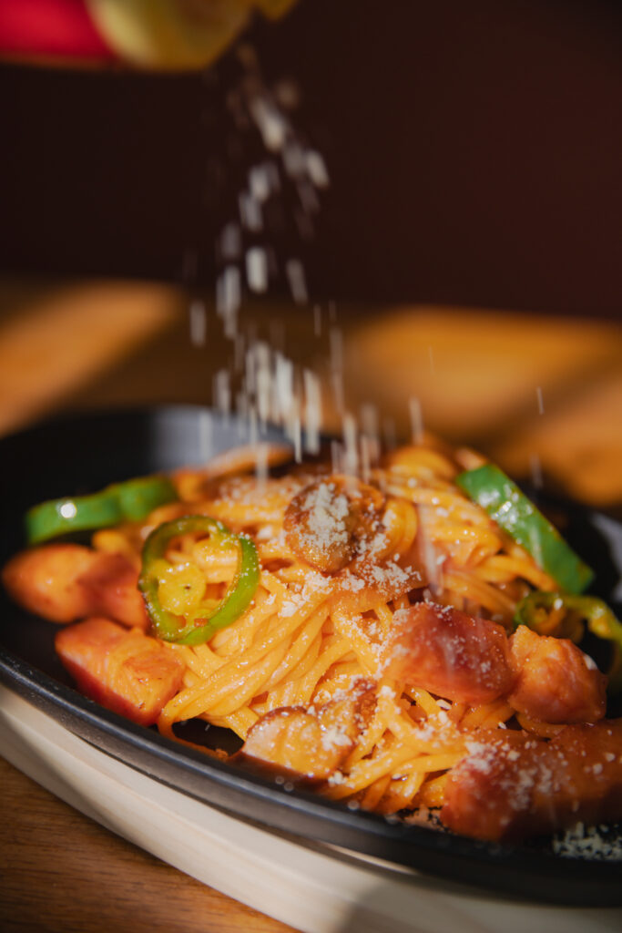 Shaking powdered parmesan cheese like falling snow over hot Japanese Napolitan pasta on a sizzling iron plate