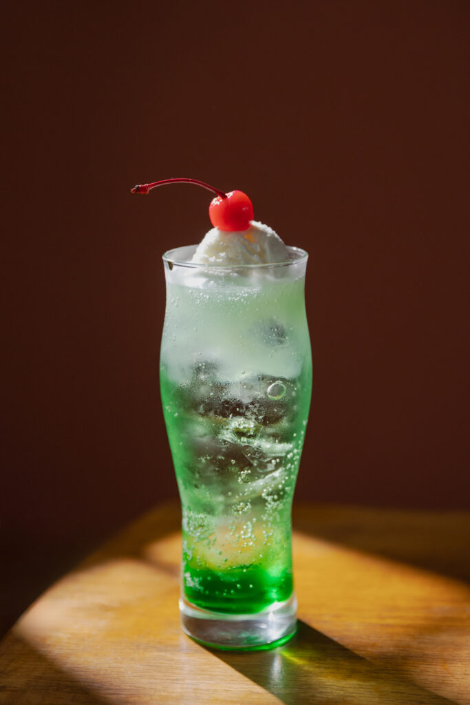 Vibrant emerald green Japanese melon soda float with a scoop of vanilla ice cream and a bright red maraschino cherry on top under dramatic studio lighting