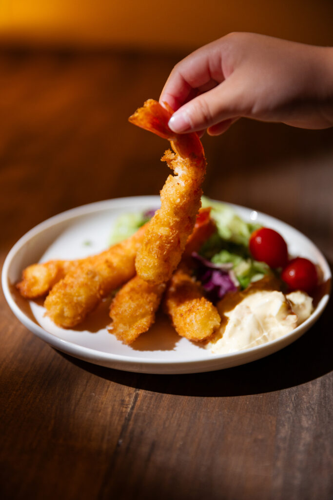 A hand lifting a single golden crispy Ebi Fry shrimp by its red tail showing incredible texture under warm light