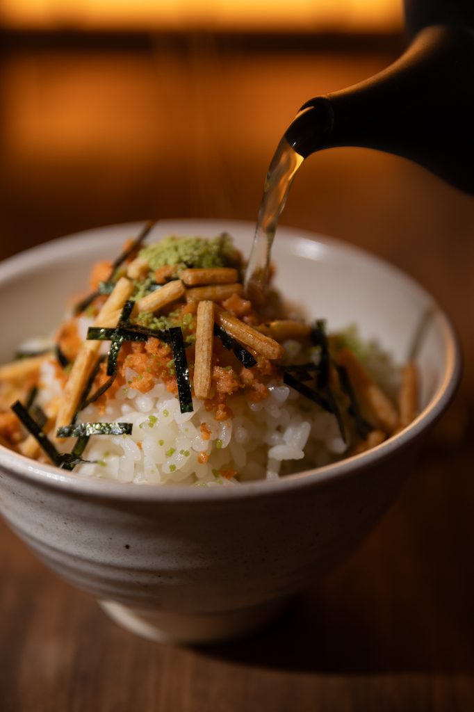 Pouring hot green tea from a black teapot onto salmon ochazuke rice bowl under warm spotlight