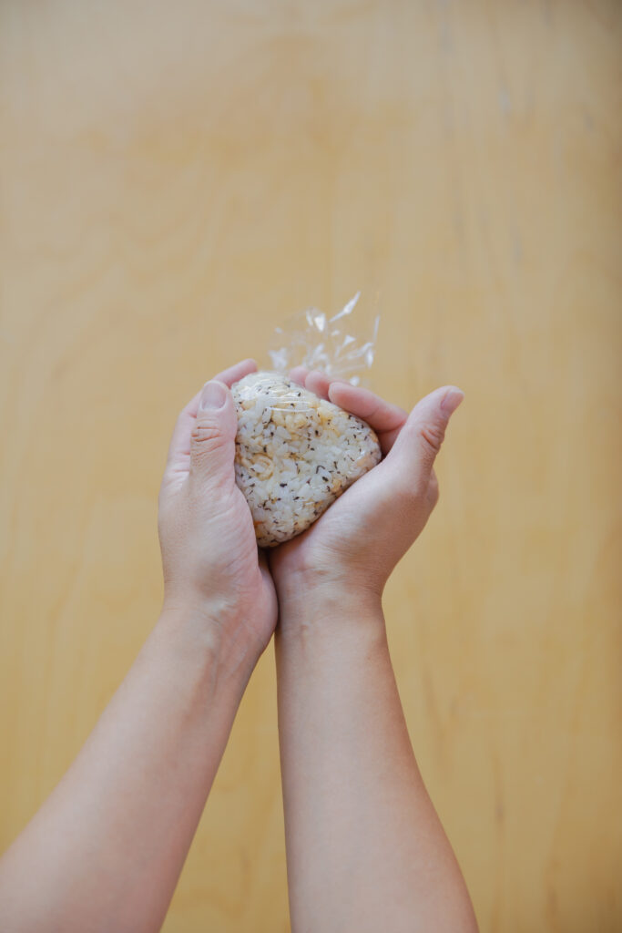 Hand-shaping a Japanese onigiri rice ball wrapped in plastic wrap over a wooden table