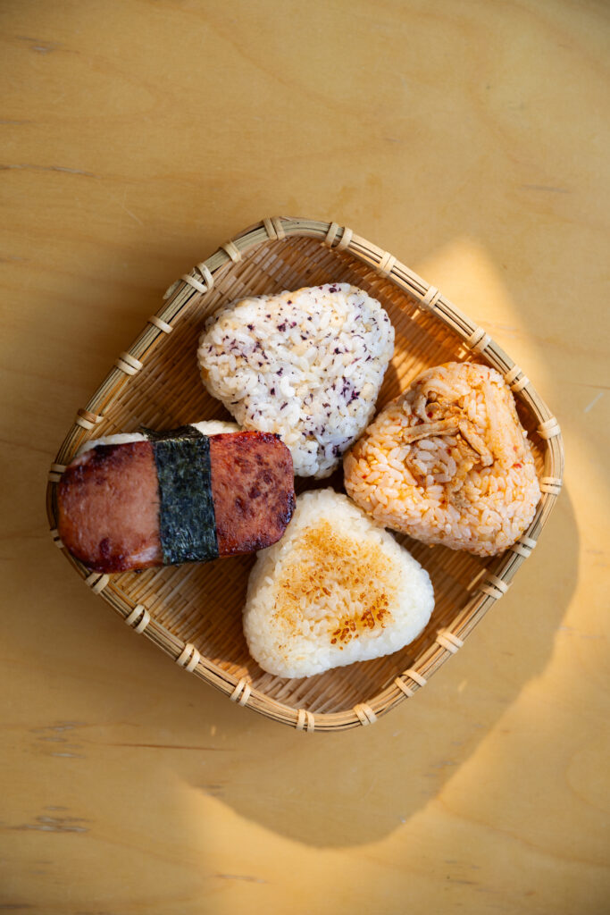 High angle view of 4 varieties of Japanese onigiri including yaki-onigiri, spam musubi, kimchi rice, and yukari shiso inside a woven bamboo basket