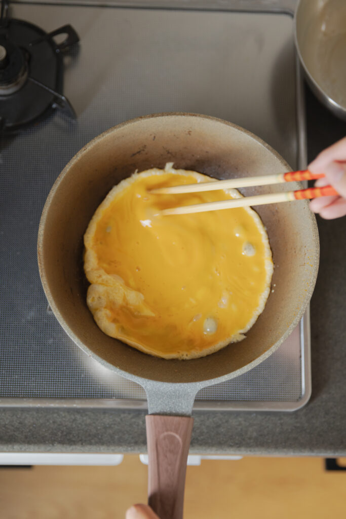 Cooking a soft-boiled fluffy egg omelet sheet in a hot frying pan with chopsticks