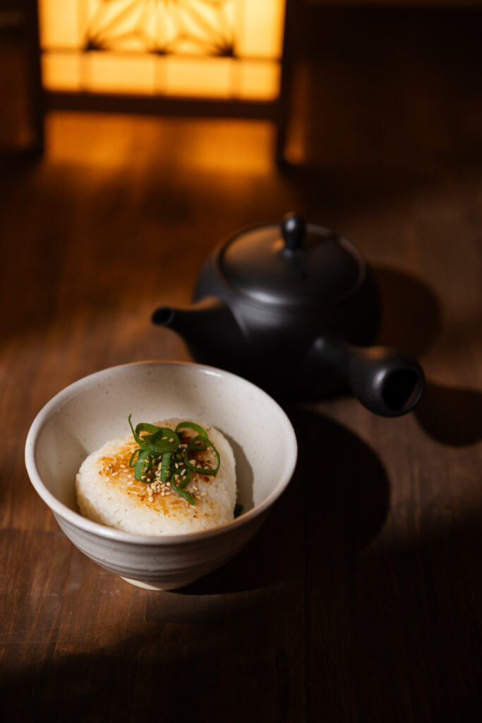 Traditional Japanese yaki-onigiri ochazuke with green onions and sesame seeds in a ceramic bowl with a black teapot behind
