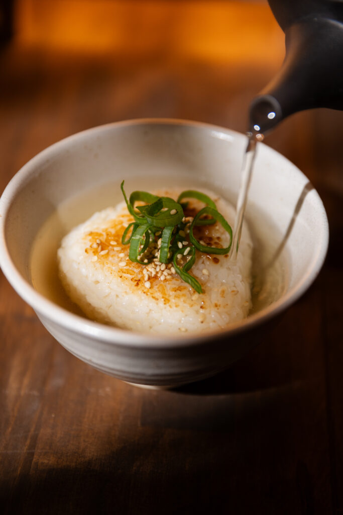 Pouring hot savory dashi broth from a black teapot onto a crispy grilled yaki-onigiri rice bowl under beautiful warm spotlight