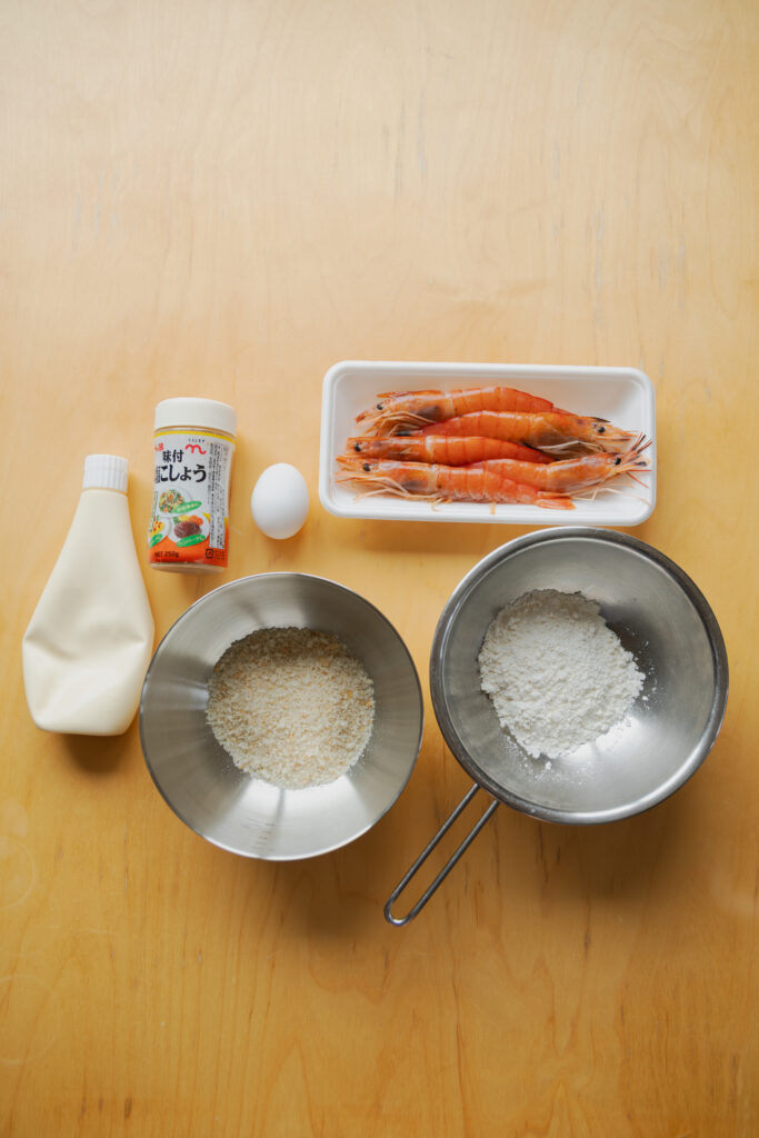 Raw fresh shrimp, bowl of panko bread crumbs, flour, egg, and mayonnaise on a light wooden table