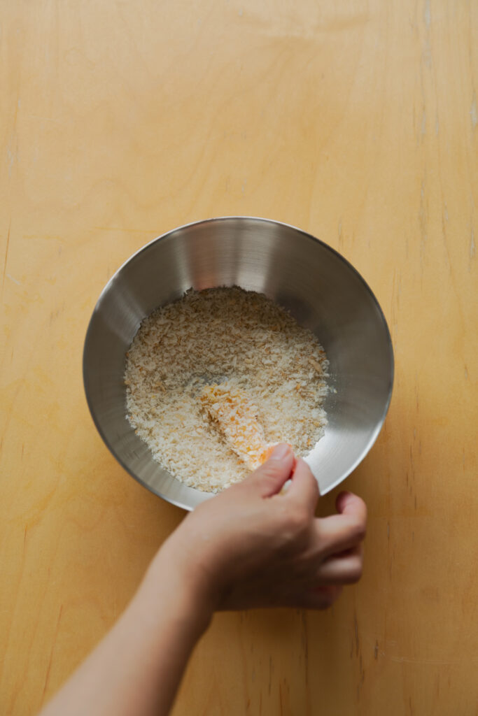 A hand coating a straight shrimp in a silver bowl filled with light, flaky Japanese Panko crumbs