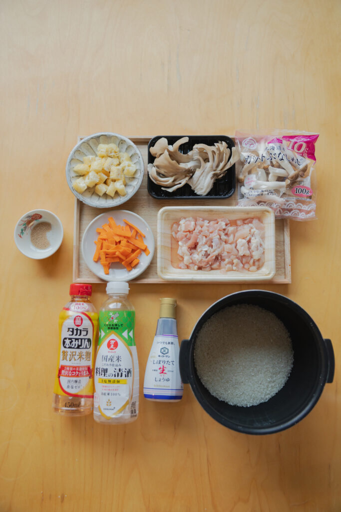 Pantry staples including raw chicken bits, maitake mushrooms, shimeji mushrooms, soy sauce, mirin, sake, and short-grain rice on a wooden tray