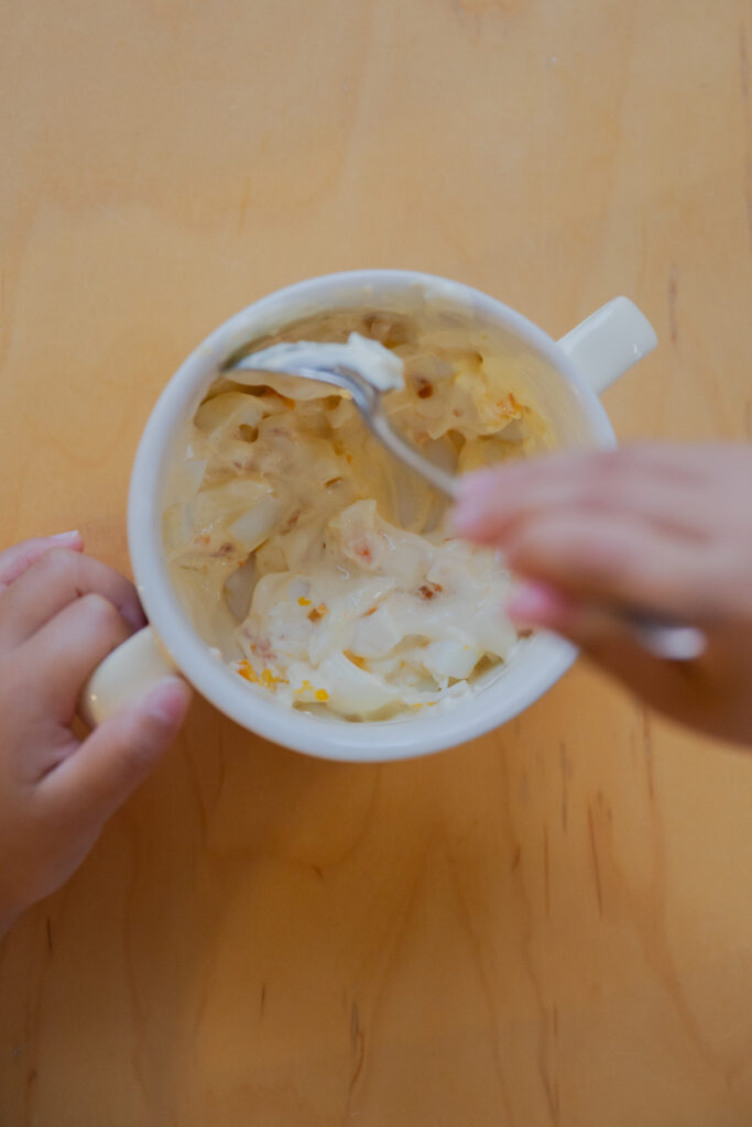 A child hands holding a double-handled white ceramic bowl and mixing a creamy egg tartar sauce with a spoon on a wooden table