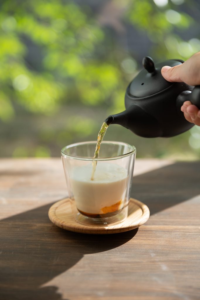 Slowly pouring hot brewed dark hojicha roasted green tea from a black ceramic Kyusu teapot into frothed milk to create elegant layers.